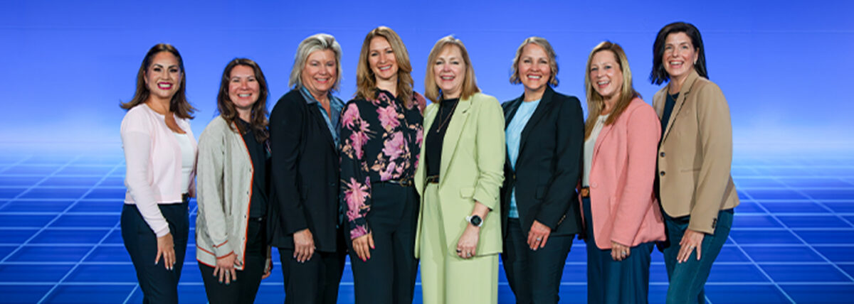 Seven women stand together and smile for a group photo against a blue backdrop during the 2026 Top Employers for Women award campaign.